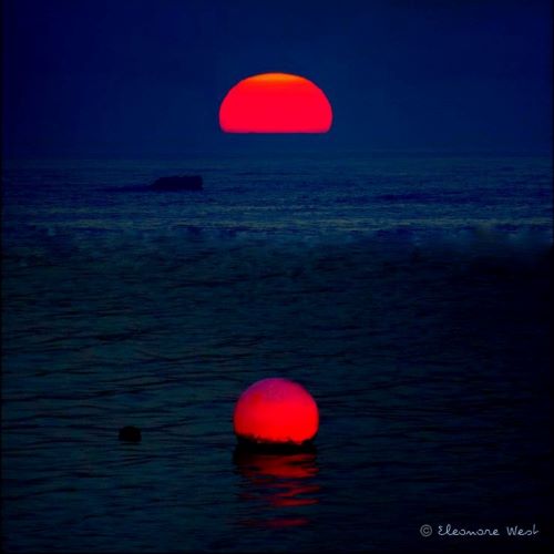 Coucher de soleil rouge sur ciel et mer bleu marine. Une bouée ronde et rouge elle aussi est placée dans l'alignement du soleil.