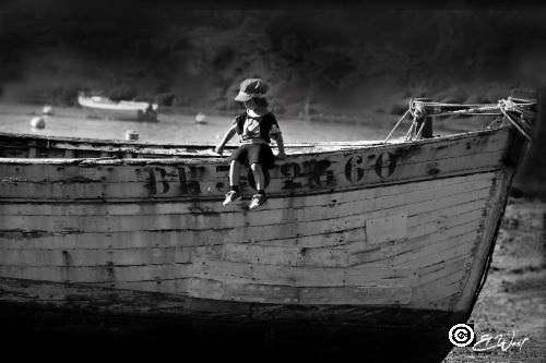 Un petit enfant, casquette sur la tête juché sur une grande barque attend la marée. Noir et blanc